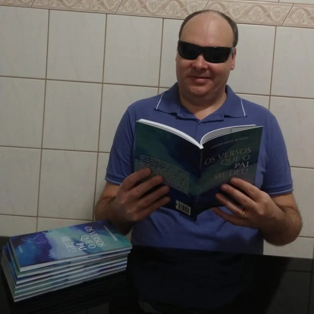 Homem de pele clara sentado segurando um livro aberto. Ele usa óculos escuros e uma camisa polo azul, com gola e punhos delineados por um filete branco. O livro que está nas mãos tem a capa em tons de azul e verde e o título em letras brancas e amarelas: “OS VERSOS QUE O PAI ME DEU"